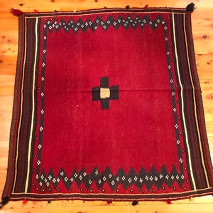 Puede incluir: Una alfombra cuadrada, tejida a mano, con un campo rojo intenso. La alfombra presenta un dise&ntilde;o central de cruz negra y beige. Un borde de rombos negros y blancos y rayas verticales en negro, beige y blanco enmarcan los bordes. Borlas adornan las esquinas.