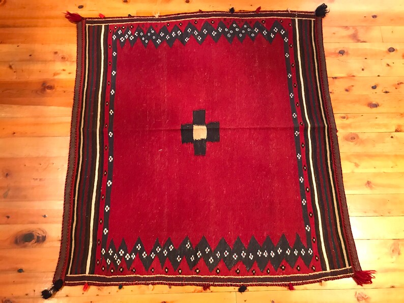 Puede incluir: Una alfombra cuadrada, tejida a mano, con un campo rojo intenso. La alfombra presenta un dise&ntilde;o central de cruz negra y beige. Un borde de rombos negros y blancos y rayas verticales en negro, beige y blanco enmarcan los bordes. Borlas adornan las esquinas.