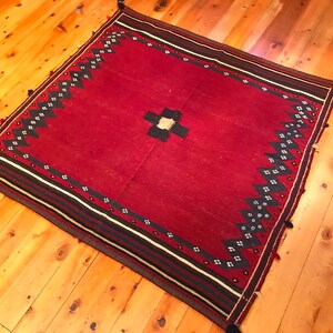 Puede incluir: Una alfombra roja cuadrada con un dise&ntilde;o central de cruz en blanco y negro. La alfombra presenta un borde con rayas negras, blancas y rojas, y un patr&oacute;n de zigzag en blanco y negro repetido. Las borlas adornan las esquinas.