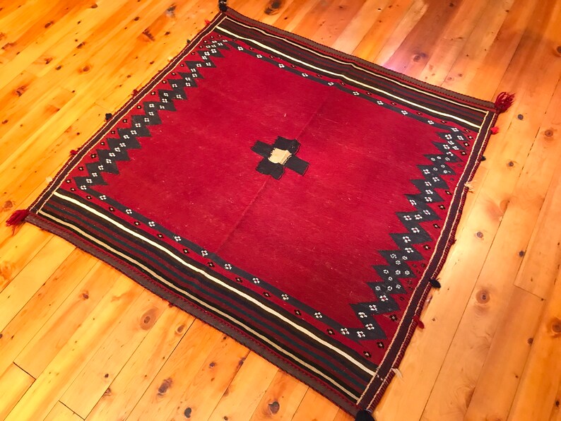 Puede incluir: Una alfombra roja cuadrada con un dise&ntilde;o central de cruz en blanco y negro. La alfombra presenta un borde con rayas negras, blancas y rojas, y un patr&oacute;n de zigzag en blanco y negro repetido. Las borlas adornan las esquinas.