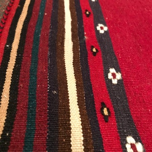 Puede incluir: Primer plano de una alfombra tejida con fondo rojo y rayas verticales en negro, beige, marr&oacute;n, verde azulado y azul marino. Una raya azul marino presenta detalles florales blancos. La alfombra tiene una superficie texturizada.