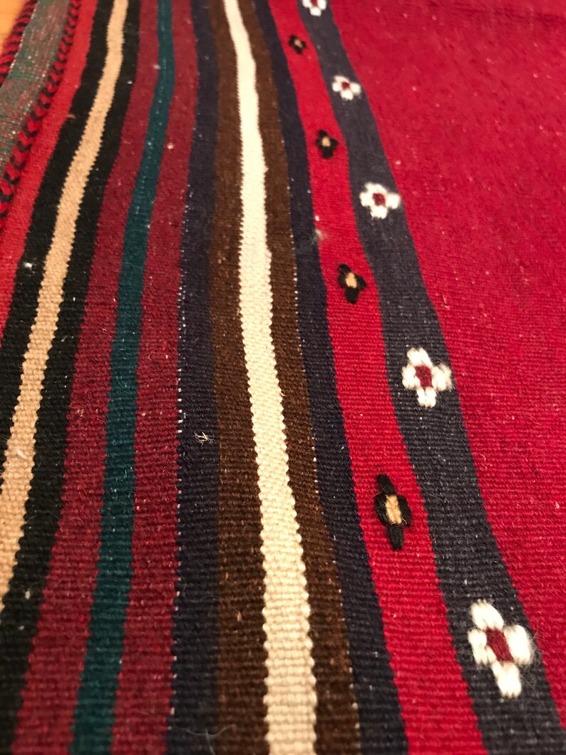 Puede incluir: Primer plano de una alfombra tejida con fondo rojo y rayas verticales en negro, beige, marr&oacute;n, verde azulado y azul marino. Una raya azul marino presenta detalles florales blancos. La alfombra tiene una superficie texturizada.