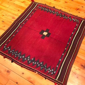 Puede incluir: Una alfombra roja cuadrada con un dise&ntilde;o de cruz central y un borde estampado. El borde presenta formas geom&eacute;tricas en blanco y negro y rayas. Las borlas adornan las esquinas. La alfombra est&aacute; colocada sobre un suelo de madera.