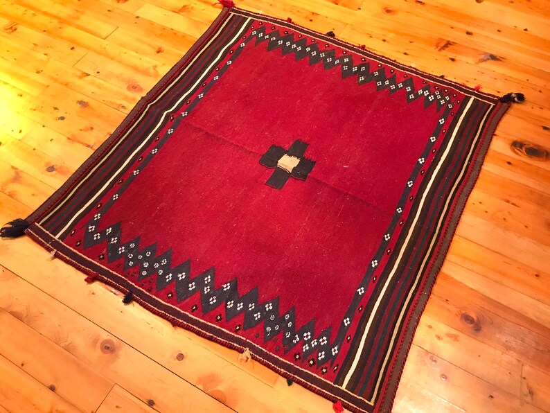 Puede incluir: Una alfombra roja cuadrada con un dise&ntilde;o de cruz central y un borde estampado. El borde presenta formas geom&eacute;tricas en blanco y negro y rayas. Las borlas adornan las esquinas. La alfombra est&aacute; colocada sobre un suelo de madera.