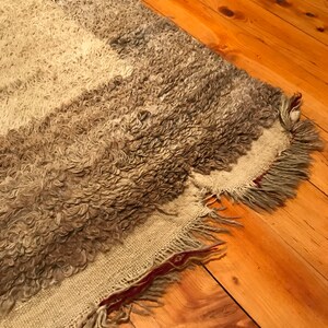 Puede incluir: Una alfombra rectangular texturizada con un pelo rizado y lanudo en tonos grises y blanquecinos. La alfombra tiene un borde con flecos y un reverso tejido. La alfombra est&aacute; colocada sobre un suelo de madera.