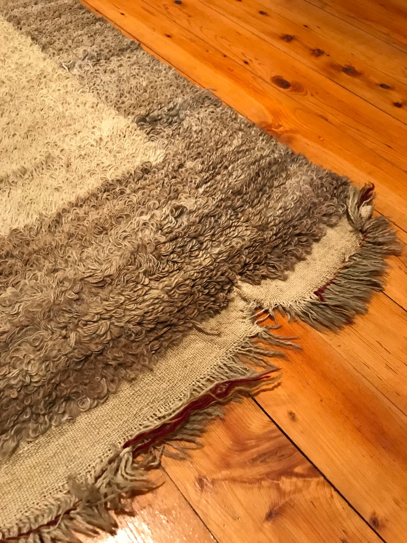 Puede incluir: Una alfombra rectangular texturizada con un pelo rizado y lanudo en tonos grises y blanquecinos. La alfombra tiene un borde con flecos y un reverso tejido. La alfombra est&aacute; colocada sobre un suelo de madera.