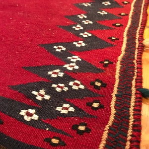 Puede incluir: Primer plano de una alfombra roja tejida con un patr&oacute;n geom&eacute;trico. La alfombra presenta un dise&ntilde;o repetitivo de tri&aacute;ngulos negros y peque&ntilde;os motivos florales blancos y negros a lo largo del borde. El borde tiene un ribete decorativo.