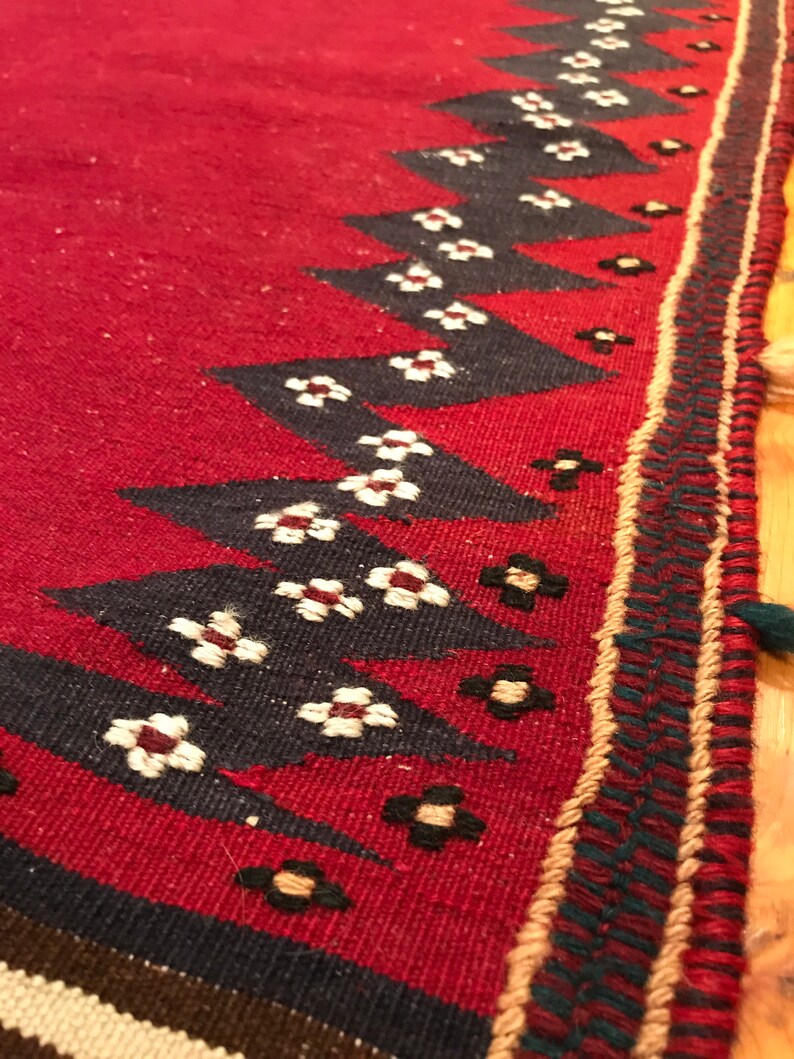 Puede incluir: Primer plano de una alfombra roja tejida con un patr&oacute;n geom&eacute;trico. La alfombra presenta un dise&ntilde;o repetitivo de tri&aacute;ngulos negros y peque&ntilde;os motivos florales blancos y negros a lo largo del borde. El borde tiene un ribete decorativo.