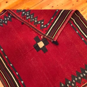 Puede incluir: Una alfombra roja plegada con un dise&ntilde;o geom&eacute;trico. La alfombra presenta un dise&ntilde;o central en forma de cruz en negro y beige, rodeado de patrones en zigzag y diamante en blanco y negro. El borde tiene rayas negras, blancas y rojas.