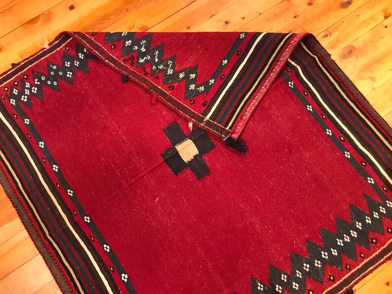 Puede incluir: Una alfombra roja plegada con un dise&ntilde;o geom&eacute;trico. La alfombra presenta un dise&ntilde;o central en forma de cruz en negro y beige, rodeado de patrones en zigzag y diamante en blanco y negro. El borde tiene rayas negras, blancas y rojas.