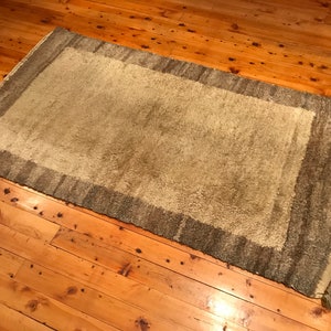 Puede incluir: Una alfombra rectangular verde claro con un borde verde oscuro y flecos. Se encuentra sobre un suelo de madera con un acabado c&aacute;lido y natural. La textura de la alfombra parece suave y afelpada, adecuada para un sal&oacute;n.