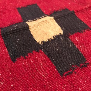 Puede incluir: Primer plano de un textil rojo tejido con un gran dise&ntilde;o de cruz negra. El centro de la cruz es de color beige. El textil tiene una apariencia texturizada con peque&ntilde;as motas blancas.