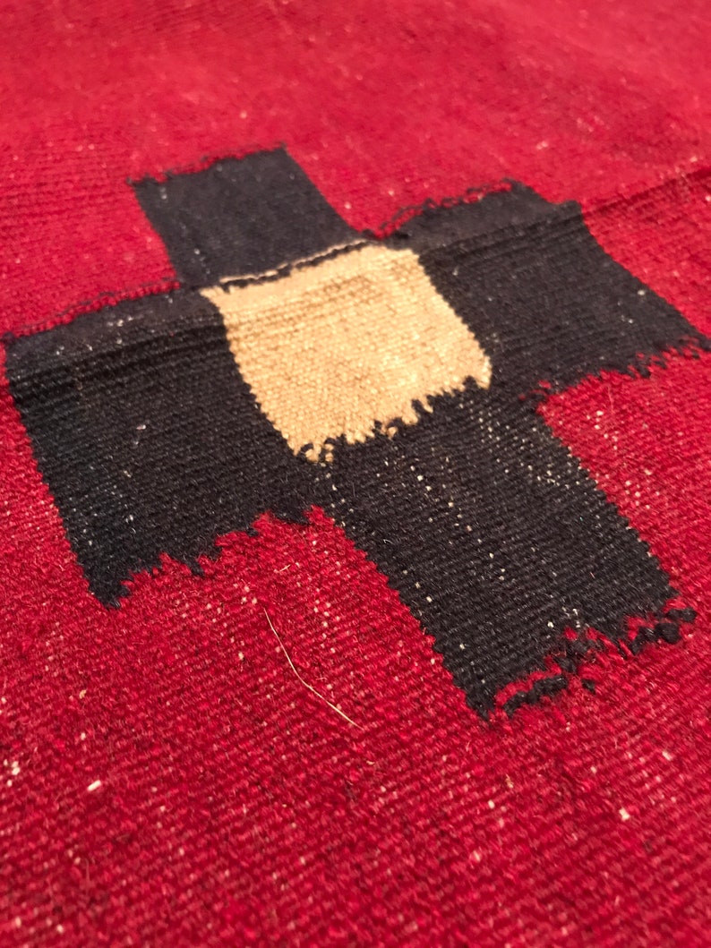 Puede incluir: Primer plano de un textil rojo tejido con un gran dise&ntilde;o de cruz negra. El centro de la cruz es de color beige. El textil tiene una apariencia texturizada con peque&ntilde;as motas blancas.