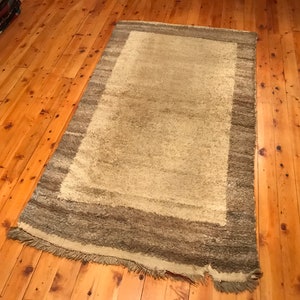 Puede incluir: Una alfombra rectangular con un centro beige claro y un borde m&aacute;s oscuro y multicolor. La alfombra tiene una textura peluda y una franja a lo largo de un borde. La alfombra est&aacute; colocada sobre un suelo de madera.