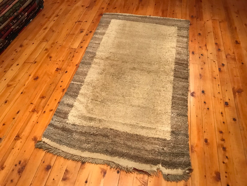 Puede incluir: Una alfombra rectangular con un centro beige claro y un borde m&aacute;s oscuro y multicolor. La alfombra tiene una textura peluda y una franja a lo largo de un borde. La alfombra est&aacute; colocada sobre un suelo de madera.