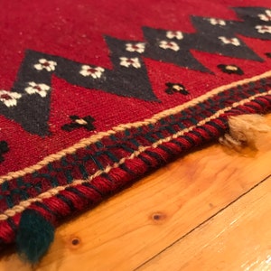 Puede incluir: Primer plano de una alfombra roja con un patr&oacute;n geom&eacute;trico. La alfombra presenta un dise&ntilde;o repetitivo de tri&aacute;ngulos negros con acentos florales blancos. El borde tiene un patr&oacute;n verde y beige. La alfombra est&aacute; sobre una superficie de madera.