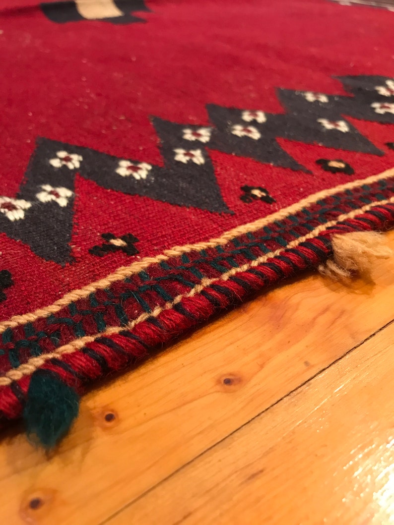 Puede incluir: Primer plano de una alfombra roja con un patr&oacute;n geom&eacute;trico. La alfombra presenta un dise&ntilde;o repetitivo de tri&aacute;ngulos negros con acentos florales blancos. El borde tiene un patr&oacute;n verde y beige. La alfombra est&aacute; sobre una superficie de madera.