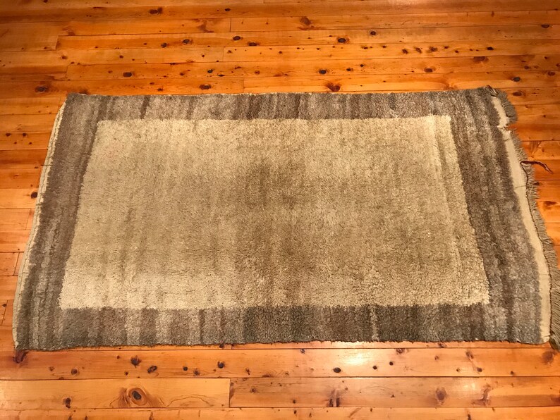 Puede incluir: Una alfombra rectangular con un centro verde claro y un borde verde m&aacute;s oscuro. La alfombra tiene una textura peluda y est&aacute; colocada sobre un suelo de madera. Los bordes de la alfombra tienen flecos.