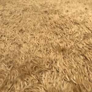 Puede incluir: Primer plano de una alfombra beige y peluda con una superficie rizada y texturizada. La alfombra tiene un aspecto suave y afelpado, con diferentes tonos de marr&oacute;n claro y beige. Las fibras est&aacute;n densamente empaquetadas, creando un aspecto c&aacute;lido y acogedor.