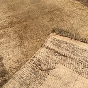 Puede incluir: Una alfombra beige con una superficie peluda y texturizada. La alfombra est&aacute; parcialmente doblada hacia atr&aacute;s, revelando un reverso tejido con un patr&oacute;n de rayas. Los bordes de la alfombra tienen flecos.
