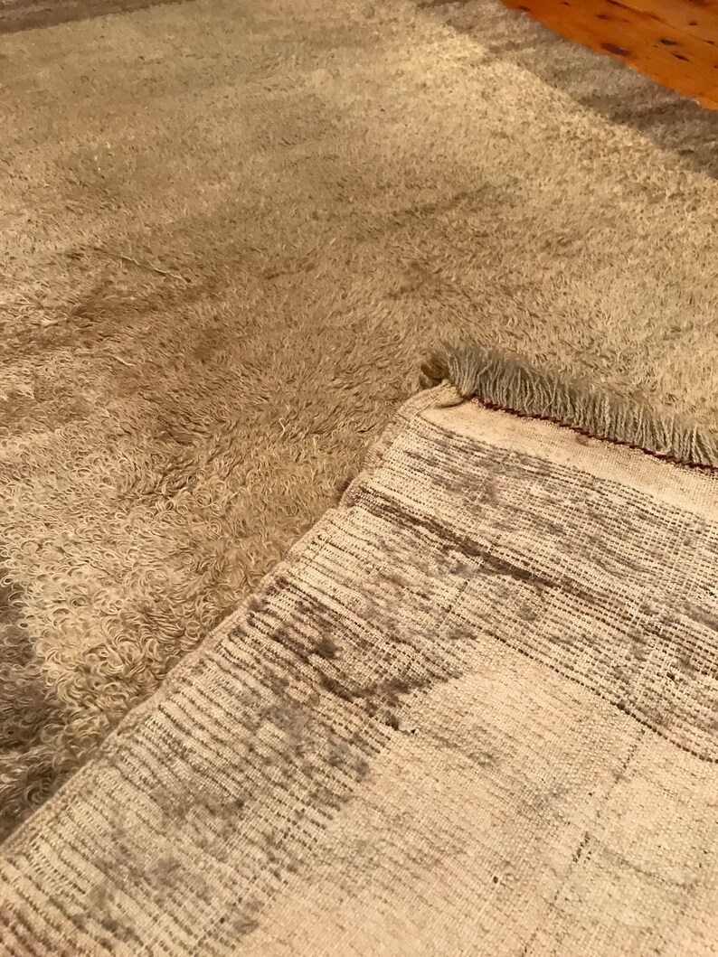 Puede incluir: Una alfombra beige con una superficie peluda y texturizada. La alfombra est&aacute; parcialmente doblada hacia atr&aacute;s, revelando un reverso tejido con un patr&oacute;n de rayas. Los bordes de la alfombra tienen flecos.