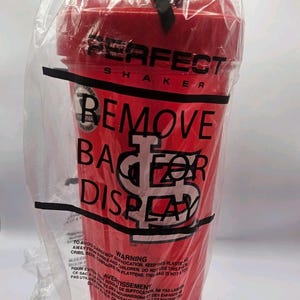 May include: A red plastic shaker bottle with a lid, enclosed in a clear plastic bag. The bottle features the text PERFECT SHAKER and a logo. The bag has black text printed on it that reads REMOVE BAG FOR DISPLAY and includes safety warnings.