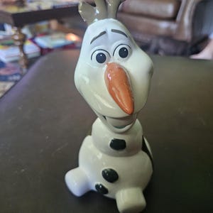 May include: A ceramic figurine of a snowman character with a white body, a bright orange carrot-shaped nose, black circular buttons on the torso, and brown twig-like hair on top. The figure has large eyes and an open mouth expression.