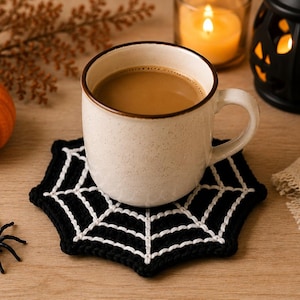 May include: A black and white crocheted spider web coaster featuring a hexagonal shape with white web detailing on a black background. The coaster supports a speckled off-white ceramic mug filled with a light brown beverage.