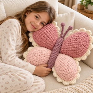 May include: A butterfly-shaped crochet pillow featuring a dark purple central body with two antennae, two upper wings in a deep rose color, and two lower wings in a lighter pink shade, all bordered by a scalloped cream-colored yarn trim.