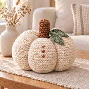 Peut inclure: Une citrouille crochetée de couleur crème avec une tige brune texturée et deux feuilles crochetées vertes. Le corps de la citrouille est orné de trois petits points de broderie en forme de flèche, de couleur rouille, alignés verticalement.