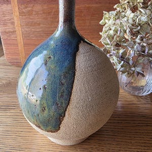 May include: A round ceramic vase with a narrow neck, featuring a dual-texture finish. One side has a smooth, glossy blue and green glaze, while the other side displays a rough, unglazed, sandy-beige clay surface with a visible granular texture.