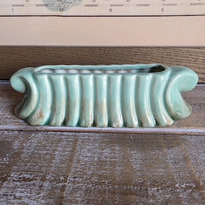 May include: A rectangular ceramic planter with a light green glaze and a vertical ribbed texture. The vessel features decorative scroll-like handles on both ends and a wide, open top, showcasing a glossy finish with subtle brown highlights.