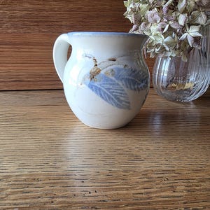 May include: A round ceramic mug with a cream-coloured base, featuring a hand-painted blue leaf design and a brown accent detail. The mug has a smooth, curved handle and a light blue rim. The surface of the ceramic has a speckled, textured finish.