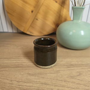 May include: A small, cylindrical ceramic cup with a dark brown, glossy glaze on the upper portion and a contrasting matte, light beige, unglazed base. The rim is slightly flared and features a smooth, rounded edge, showcasing a simple design.