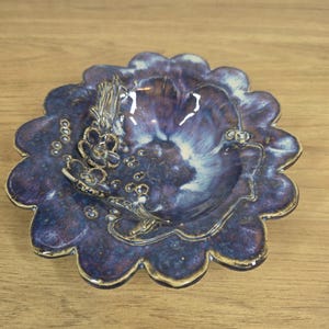 May include: A round ceramic dish with a scalloped edge, featuring a deep purple and blue glaze with white highlights. The interior surface is decorated with raised, sculpted floral elements and small circular accents, creating a textured appearance.