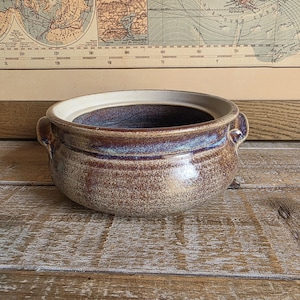 May include: A round ceramic bowl with two small side handles, featuring a speckled brown and tan exterior glaze that transitions into a deep blue and purple band near the rim, with a smooth, light-coloured interior surface and a wide opening.