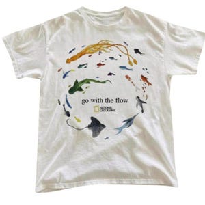 Può includere: Una t-shirt bianca a maniche corte con una disposizione circolare di varie illustrazioni colorate di vita marina, tra cui una razza e un calamaro. Il testo "go with the flow" e il logo National Geographic sono stampati al centro.