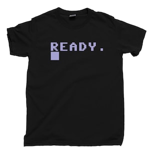 Könnte beinhalten: Ein schwarzes T-Shirt mit kurzen Ärmeln und dem verpixelten Text "READY." in Hellviolett, ergänzt durch ein kleines hellviolettes Quadrat direkt unter dem Buchstaben R, das eine klassische Computer-Befehlszeilenschnittstelle imitiert.