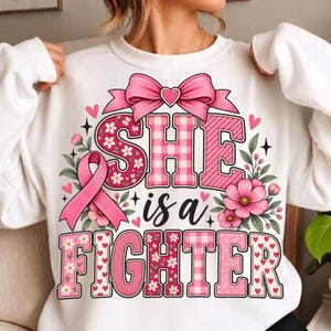 Op de afbeelding: Een wit sweatshirt met een roze grafisch ontwerp met de tekst "SHE is a FIGHTER". Het ontwerp bevat een roze lint, een grote roze strik en bloemmotieven in roze tinten. De letters zijn gevuld met ruitjes, strepen en bloemmotieven.