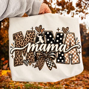 Puede incluir: Una sudadera blanca con la palabra MAMA en letras grandes con patrones variados que incluyen estampado de leopardo, lunares blancos y negros, y un tablero de ajedrez marrón. Un mama en cursiva se superpone, con un lazo y corazones.