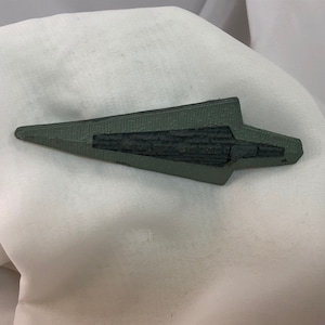 Puede incluir: Un objeto gris en forma de punta de flecha con un centro oscuro y texturizado. El objeto tiene un diseño geométrico y está sobre un fondo blanco. La punta de flecha es probablemente un objeto decorativo o una obra de arte.