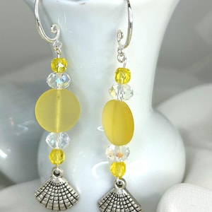May include: Dangle earrings featuring silver-toned metal hooks, clear faceted crystal beads, small yellow round beads, and a larger matte yellow circular disc. The design concludes with a silver-toned metal charm shaped like a seashell fan.