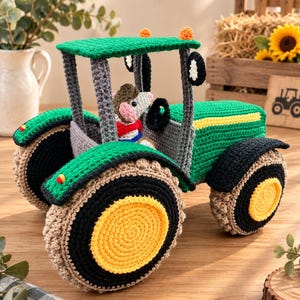 Op de afbeelding: Een gehaakte speelgoedtractor in groen, geel en zwart met een klein gehaakt dierenfiguurtje op de bestuurdersstoel. De tractor heeft een groen dak, grijze steunpilaren, zwarte spatborden en grote geel-zwarte wielen met beige accenten.