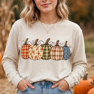 May include: A long-sleeve cream-colored sweatshirt featuring a graphic print of five pumpkins in a row. Each pumpkin displays a unique pattern, including plaid, floral, and gingham designs in shades of orange, green, blue, and brown.