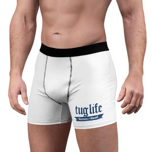 May include: White boxer briefs featuring a black waistband and black stitching along the front panel. The lower right side displays the blue text "tug life" in a stylised font, with the words "Restore the Hood" printed underneath.