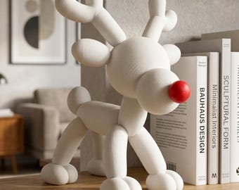 May include: A matte white reindeer figurine with a smooth, rounded, balloon-like aesthetic. The sculpture features a prominent, spherical, bright red nose, upright antlers, and four sturdy legs, standing on a wooden shelf next to several books.