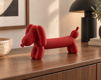 May include: A matte red sculpture shaped like a balloon animal dachshund. The figure features a long body, four short legs, a rounded snout, floppy ears, and a small tail, all rendered in a smooth, solid red finish with a balloon-like aesthetic.