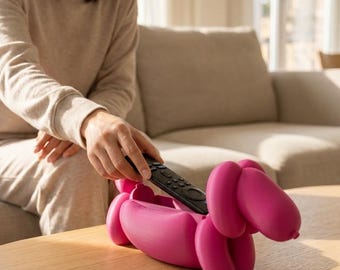 May include: A bright pink, hollow, balloon-style dog figurine designed as a storage container. The dog shape features rounded, inflated-looking segments and a smooth matte finish. A black television remote control is placed inside the open top.