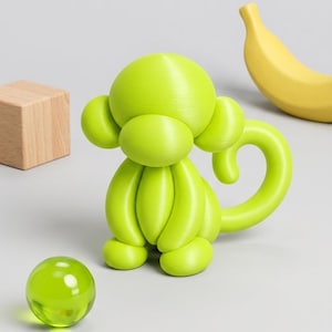 Puede incluir: Una figura de mono de color verde lima con una estética suave y redondeada similar a un globo, que presenta una cola enroscada, orejas grandes y una cara ovalada prominente. El mono está colocado cerca de una pequeña esfera de vidrio verde.