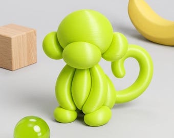 May include: A lime green monkey figurine with a smooth, rounded balloon-like aesthetic, featuring a curled tail, large ears, and a prominent oval-shaped face. The monkey is positioned near a small, translucent green glass sphere, a wooden block, and a yellow banana.